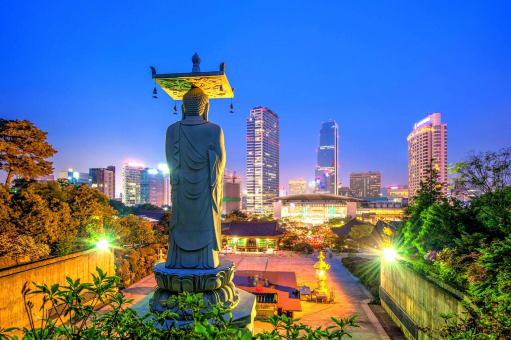 Large Buddha statue at a temple overlooking a modern Asian city skyline at dusk